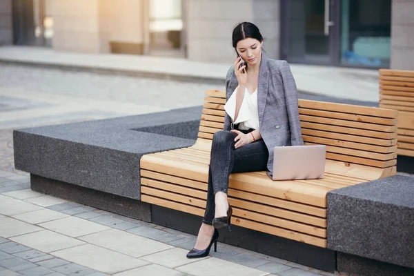 Zakenvrouw in een zakenpak zittend op tafel en pratend op een smartphone kijkend naar het laptopscherm en genietend van een pauze — Stockfoto