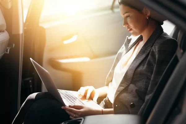Podnikatelka v bundě sedí v autě na zadním sedadle a pracuje na notebooku a kontroluje pracovní poštu — Stock fotografie