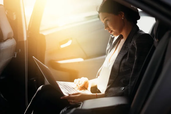 Podnikatelka v bundě sedí v autě na zadním sedadle a pracuje na notebooku a kontroluje pracovní poštu — Stock fotografie