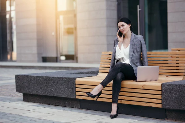 Mooie zakenvrouw in een jasje zittend op een bankje op straat en pratend op de telefoon en post laptop — Stockfoto