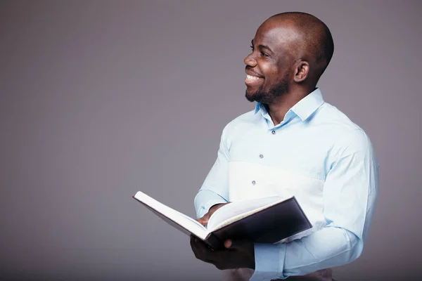 Afrykański pracownik biurowy w białej koszuli, przechodzący przez grubą książkę i uśmiechnięty. Przeglądanie i zatwierdzanie materiałów drukowanych. — Zdjęcie stockowe