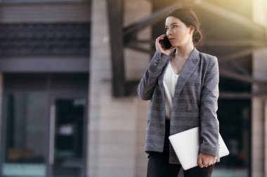 Elinde dizüstü bilgisayarla telefonda konuşan ceketli güzel bir iş kadınının sokak fotoğrafı.