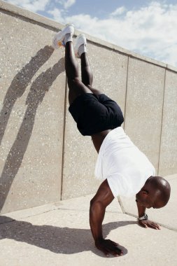 Koyu tenli bir sporcu elleri yerde, ayakları beton bir duvarda dinleniyor. Parkta sabah egzersizleri.