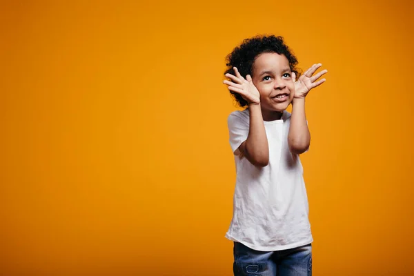 Menino afro-americano em uma camiseta branca segura as mãos perto de seu rosto e tolos em torno de um fundo laranja . — Fotografia de Stock