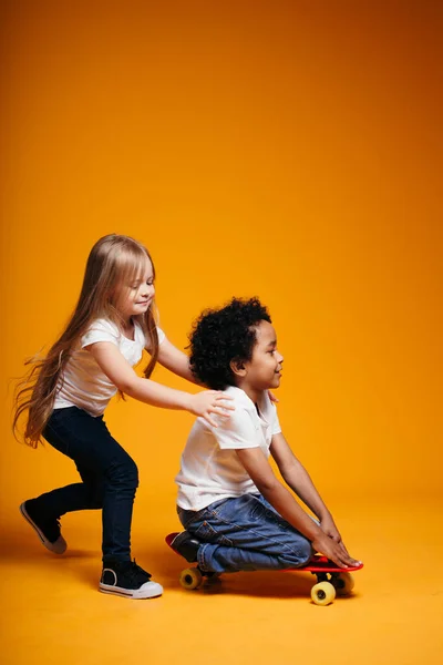 Blond meisje rijdt een krullend zwart jongen op een skateboard op een oranje achtergrond in de studio. — Stockfoto