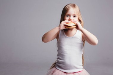 Etekli ve kolsuz tişörtlü küçük sarışın kız büyük bir hamburger yiyor ve arka plandaki kameraya bakıyor.