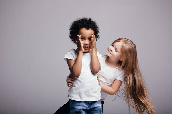 Little long-haired girl stands behind a curly African-American boy who covers his face with his hands on a gray background