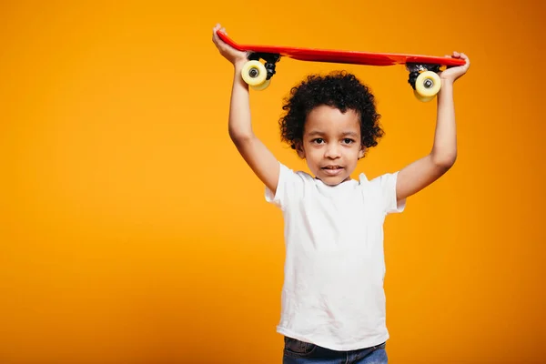 白いTシャツの子供の男の子はオレンジの背景に彼の頭の上に赤いスケートボードを保持しています — ストック写真