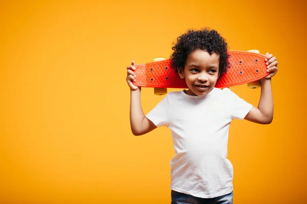 Zwart jongen kind in een wit T-shirt houdt een skate op zijn schouders op een oranje achtergrond in de studio — Stockfoto