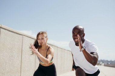 Beyaz tişörtlü profesyonel bir esmer koşu antrenörü sarışın öğrencisine bir süre sokakta nasıl koşulacağını öğretiyor.