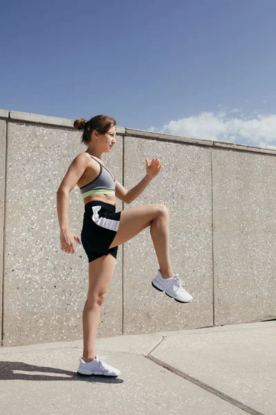Una deportista en ropa deportiva está haciendo un ejercicio para levantar las piernas para balancear los músculos de los cuádriceps y las nalgas en la calle . — Foto de Stock