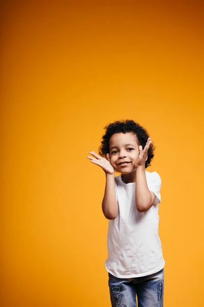 Kinky African American Boy em uma camiseta branca e jeans segura as mãos pelo rosto e olha para a câmera em um fundo laranja no estúdio — Fotografia de Stock