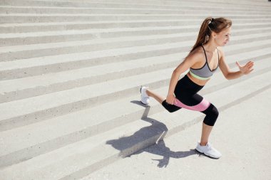 Pembe lastik spor bantlı pilates eğitmeni parkta göğsüne yakın el ele tutuşarak çömeliyor..