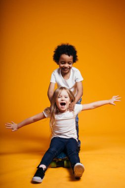 Siyah tenli kıvırcık çocuk kollarını kaykayın kenarlarına doğru açan Avrupalı bir kıza biniyor.