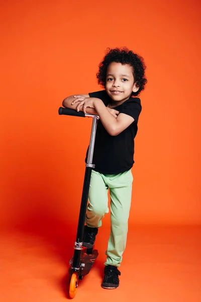 Retrato completo de um menino afro-americano montando uma scooter em um estúdio em um fundo laranja — Fotografia de Stock