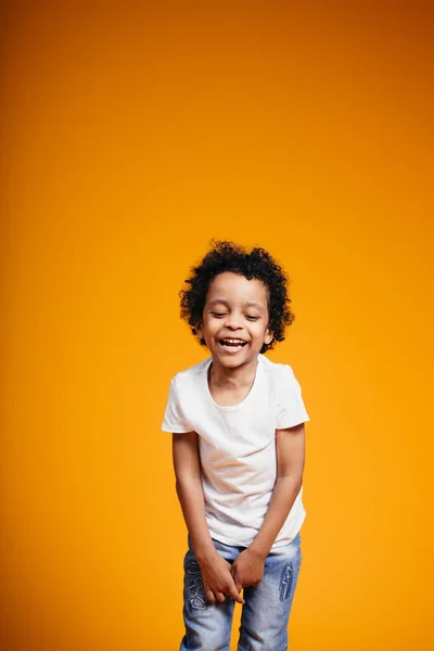 Garçon éthiopien aux cheveux bouclés dans un T-shirt blanc redresse les jeans, rit et ferme les yeux sur un fond orange dans le studio — Photo