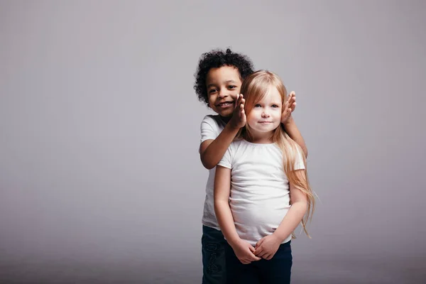 Retrato de dos niños de diferentes nacionalidades de pie juntos. El chico cierra las orejas de la chica sobre un fondo claro . —  Fotos de Stock