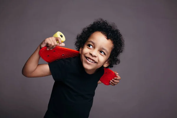 Close-up retrato de um menino afro-americano encaracolado em uma camiseta preta segurando skate vermelho nas costas e sorrindo para a câmera — Fotografia de Stock