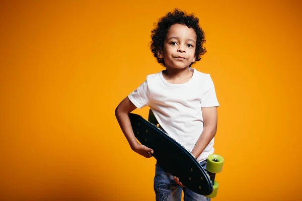 Kinky Maracanian Boy dans un T-shirt blanc et un jean étreint une planche à roulettes et l'appuie sur sa poitrine. Photographie studio d'enfants — Photo