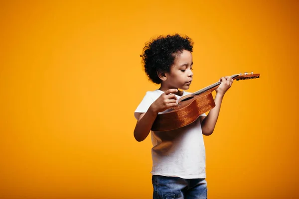 Menino em uma camiseta branca segura uma guitarra ukulele perto de seu peito e olha atentamente para cordas contra um fundo laranja — Fotografia de Stock