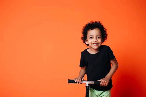 Retrato de um menino caracolado rindo alegremente montando uma scooter em um fundo laranja . — Fotografia de Stock