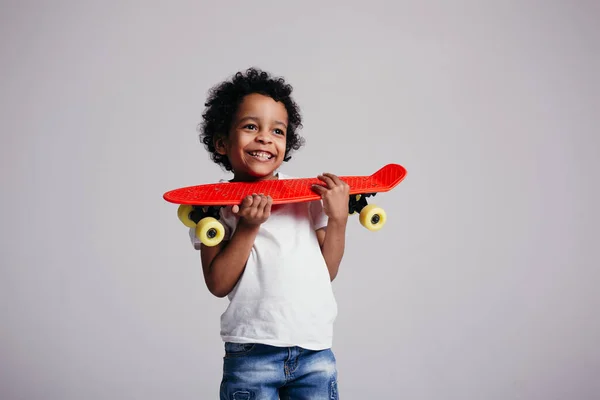 Een zwarte krullende jongen drukt een rood longboard voor zichzelf en glimlacht naar de camera in de studio op een lichte achtergrond — Stockfoto