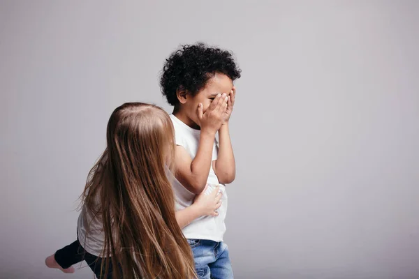 De donkere jongen sloot zijn ogen en overweegt om verstoppertje te spelen met een meisje met blond haar op een grijze achtergrond.. — Stockfoto