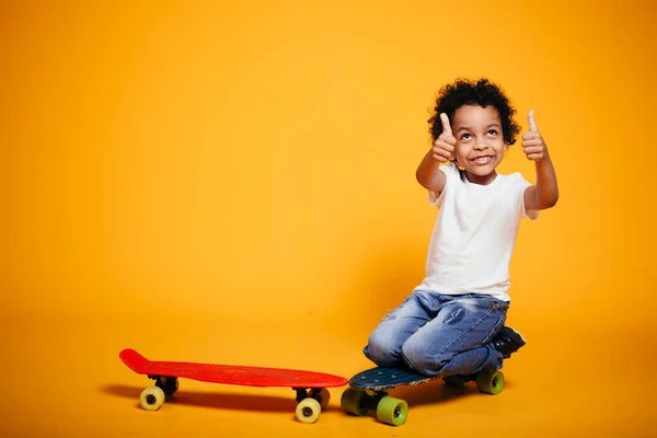 Liten pojke i vit T-shirt sitter på knä på en gul bakgrund och visar två tummar upp — Stockfoto