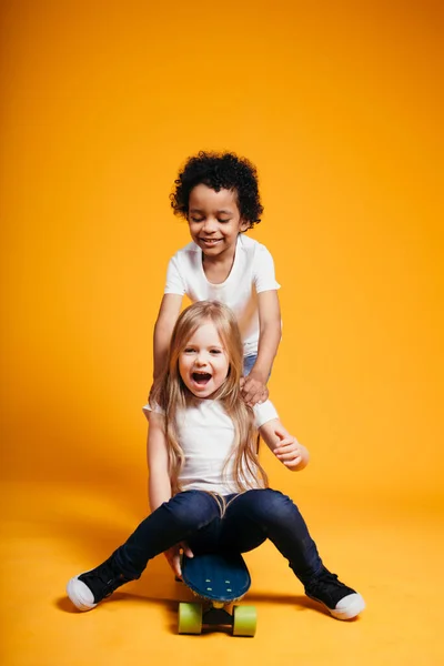 Krullende donkere huid jongen rijdt zijn kleine vriendin op een longboard, en het meisje lacht op een oranje achtergrond — Stockfoto
