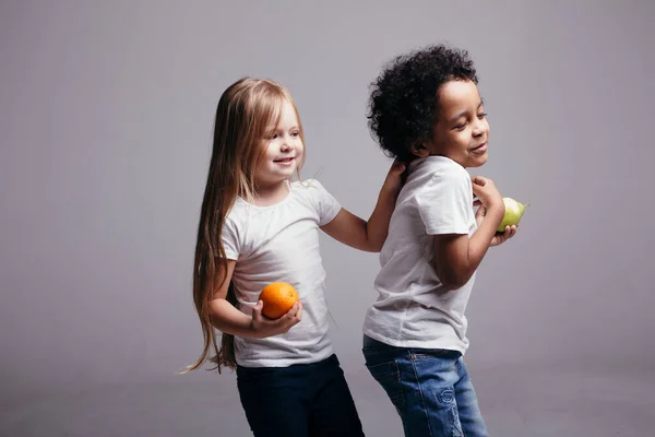 Een klein meisje kietelt de nek van haar vriend en ze houden een sinaasappel en een appel in hun handen op een lichte achtergrond. — Stockfoto