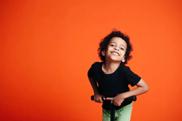 Um menino de cabelos encaracolados em uma camiseta preta monta uma scooter e brinca se inclina para a frente em direção à câmera no estúdio — Fotografia de Stock