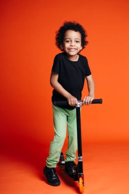 Turuncu arka planda scooter süren küçük siyah bir çocuğun tam boy portresi.