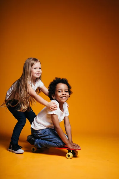 Menina loira monta um menino negro em um longboard em um fundo laranja no estúdio — Fotografia de Stock