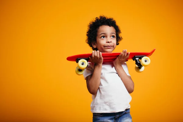 Un garçon à la peau foncée et souriant tient un longboard rouge dans ses mains et le presse sur sa poitrine — Photo