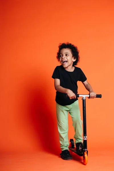 Retrato completo de um menino etíope rindo e de pé em uma scooter em um fundo laranja no estúdio — Fotografia de Stock