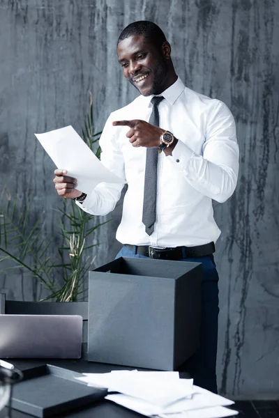 Ciemnoskóry pracownik firmy uśmiecha się, trzyma kawałek papieru w ręku i wskazuje palcem na niego w biurze — Zdjęcie stockowe