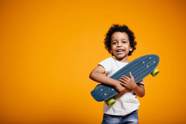 Ethiopian Boy in een wit T-shirt en jeans knuffelt een skateboard en drukt het tegen zijn borst op oranje achtergrond. — Stockfoto