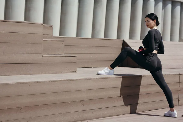 Pilates entrenador en un chándal negro estira los músculos internos de las caderas con su pie en un banco del parque —  Fotos de Stock