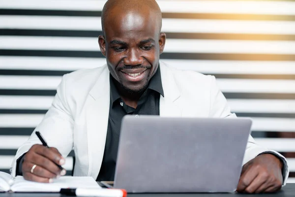 Donkere directeur van het bedrijf in wit pak kijkt aandachtig door rapporten voor het half jaar op laptop zitten aan een tafel — Stockfoto