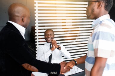 Siyahi iş ortakları el sıkışıyor ve arka planda neşeli bir yönetici el sıkışıyor ve gülümsüyor.