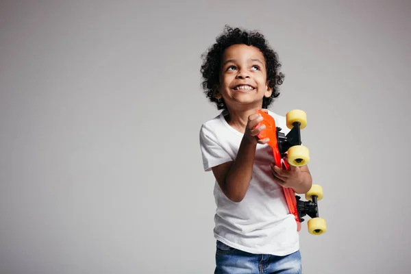 Een donkergekleurde krullende jongen in een wit T-shirt regisseerde een rood longboard in de camera en kijkt op en glimlacht op een witte achtergrond — Stockfoto