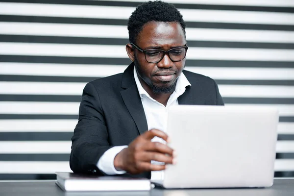 Afroamerický podnikatel v černém kostýmu a brýlích sedí za stolem a začíná pracovat na projektu na notebooku. — Stock fotografie