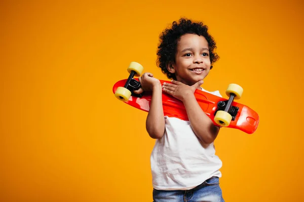 Un garçon à la peau foncée dans un T-shirt blanc presse un longboard rouge sur sa poitrine sur un fond orange dans le studio et sourit — Photo