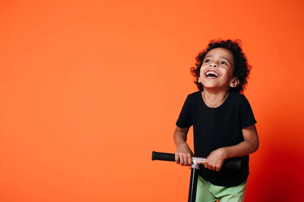 Retrato de um menino afro-americano que joga a cabeça para trás e montando uma scooter em um estúdio em um fundo laranja . — Fotografia de Stock