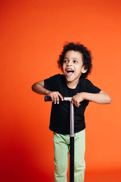 Retrato completo de um menino negro de pele encaracolada rindo e montando uma scooter em um estúdio em um fundo laranja — Fotografia de Stock