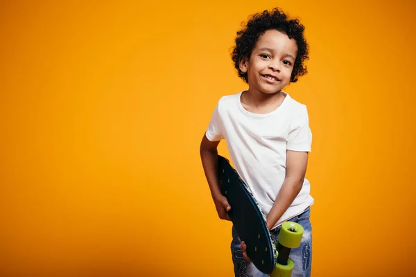Menino marroquino em uma camiseta branca segura um skate e olha para a câmera sorrindo em um fundo laranja — Fotografia de Stock