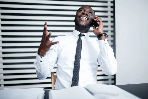 Μαύρος μάνατζερ με λευκό πουκάμισο χαμογελάει και γελάει μιλώντας στο τηλέφωνο με επενδυτές και λύνει εργασιακά θέματα στο γραφείο — Φωτογραφία Αρχείου