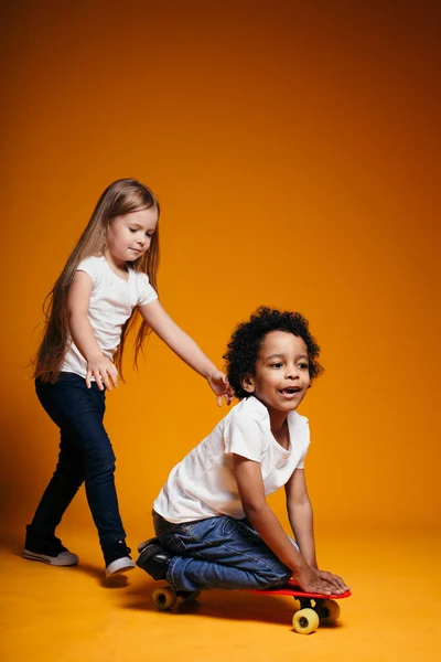 Blond meisje lacht luid terwijl rollen haar zwarte vriend op een skateboard op een oranje achtergrond in de studio — Stockfoto