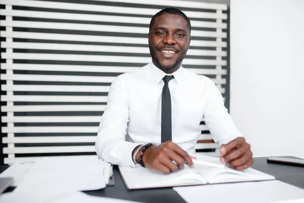 Portret firmy czarny uśmiechnięty biznesmen w białej koszuli i krawacie siedzący w biurze przy stole z dokumentami — Zdjęcie stockowe