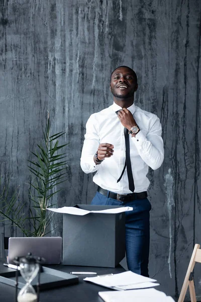 Um homem de pele escura com uma camisa branca endireita sua gravata e sorri enquanto olha para a câmera no escritório . — Fotografia de Stock